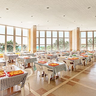 На фото: часть помещения обеденной зоны столовой, большие панорамные окна с видом на море, обеденные столы на четырех человек, на столах подносы с фруктами, помещение оборудовано видеокамерами, потолки - подвесные, светильники - точечные, полы - гранитная крошка