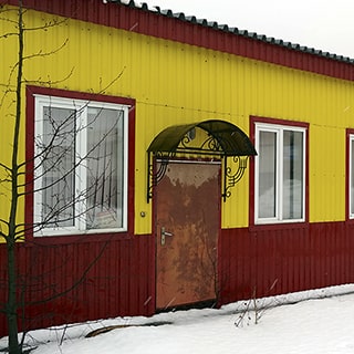 На зимнем фото: часть фасада нежилого одноэтажного здания, облицовка фасада и кровля выполнены из металлического профилированного листа, окна - стеклопакеты, входная дверь - металлическая под защитным козырьком, территория перед фасадом покрыта снегом